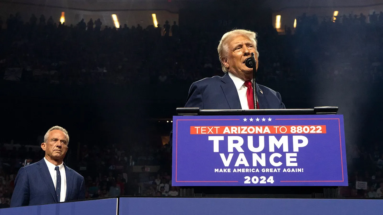 دونالد ترامب يتحدث في تجمع انتخابي بينما يقف روبرت كينيدي الابن خلفه، مع لافتة تدعو لدعمهما في الانتخابات.