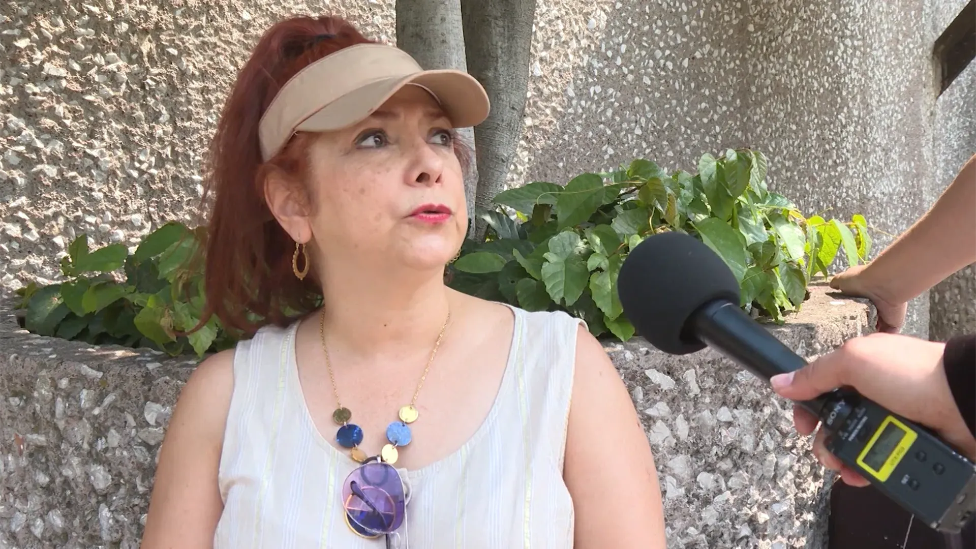 امرأة تتحدث إلى صحفي، مرتدية قبعة وقميصًا أبيض، بينما تظهر خلفها نباتات خضراء وجدار رمادي، تعبيرها يدل على الجدية.