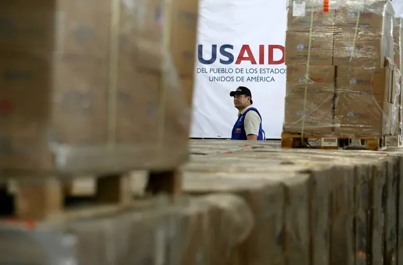 موظف في وكالة التنمية الدولية الأمريكية (USAID) يقف بين صناديق المساعدات الغذائية، مع خلفية تشير إلى تجميد المساعدات الإنسانية.