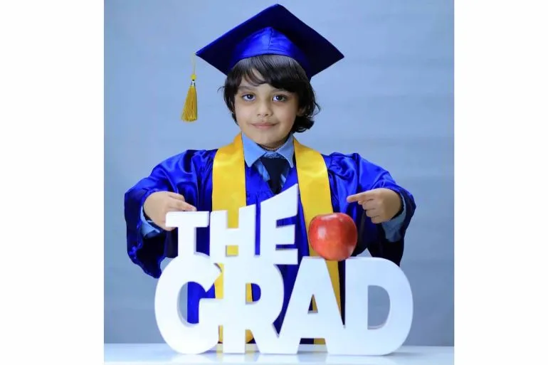 طفل يرتدي قبعة التخرج وزيّاً أكاديمياً، يشير بفخر إلى كلمة "THE GRAD" مع تفاحة حمراء، معبراً عن فرحة التخرج والإنجاز.
