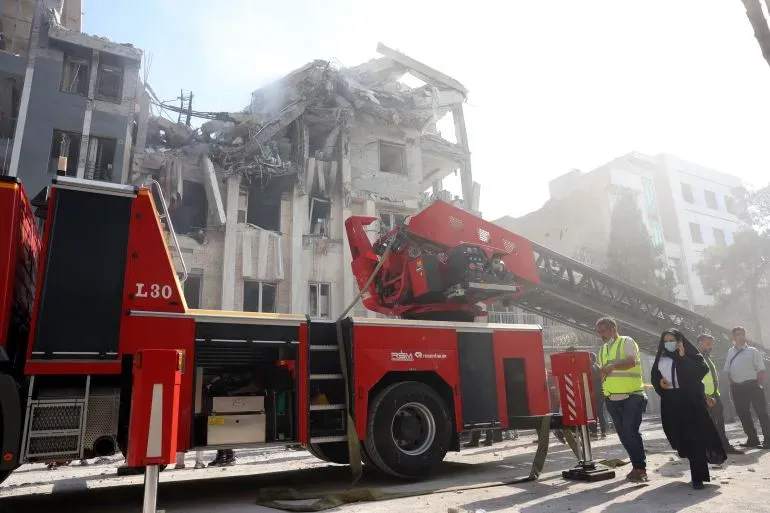 مركبة إطفاء تهرع إلى موقع بناء متضرر، حيث يظهر الدمار الناتج عن الهجوم الإسرائيلي على إيران، مع وجود أشخاص يتفقدون الأضرار.