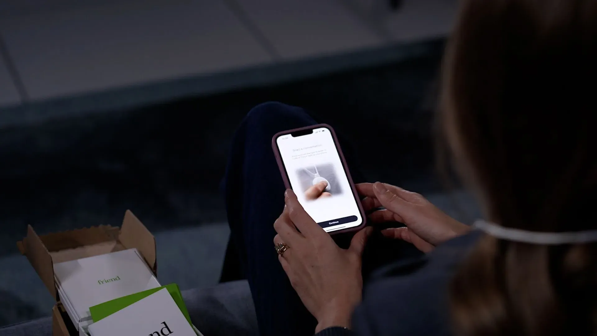 امرأة تحمل هاتفًا ذكيًا وتستخدم تطبيقًا لرفيق الذكاء الاصطناعي "Friend"، مع تفاصيل حول المنتج في الخلفية.