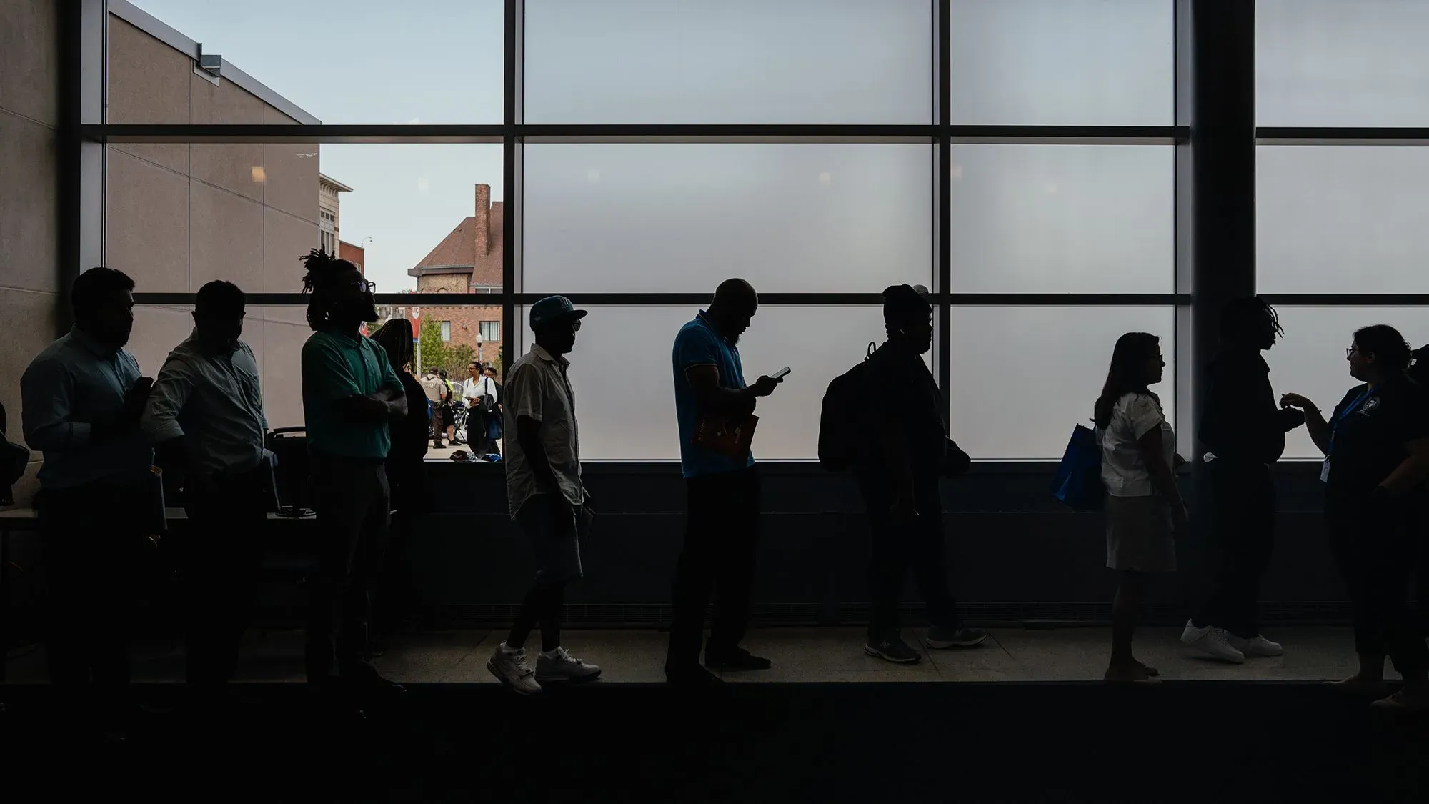 طابور من الأشخاص ينتظرون في مكان داخلي، مع إضاءة خلفية تبرز ظلالهم، مما يعكس تأثيرات سوق العمل والبطالة في الولايات المتحدة.