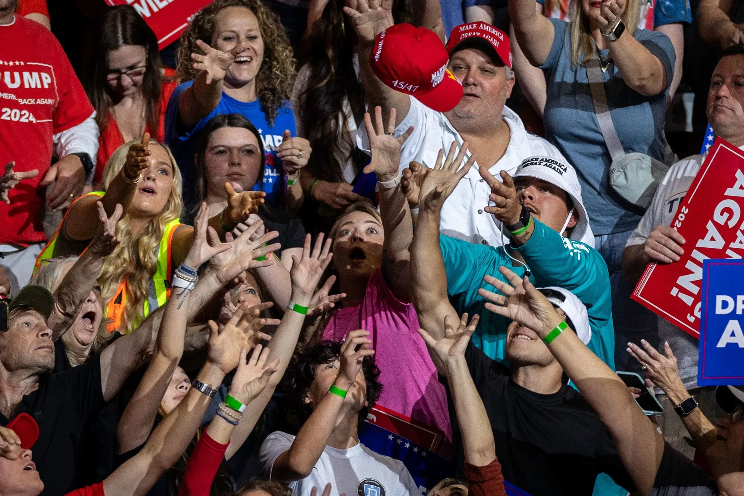 حشد من المؤيدين في تجمع سياسي، يمدّون أيديهم نحو قبعة تحمل شعار ترامب، مع لافتات تدعم حملته الانتخابية لعام 2024.