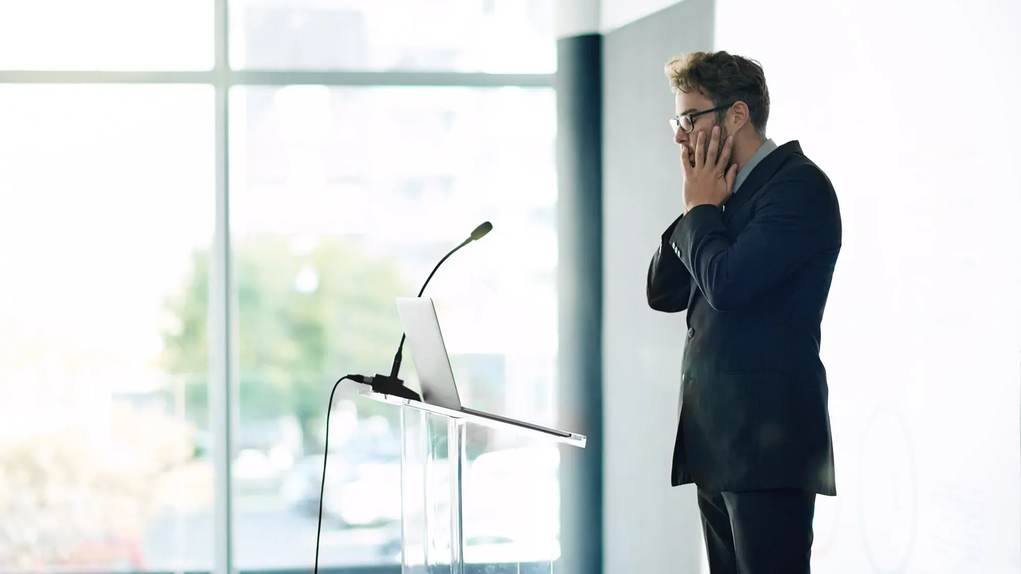 رجل يرتدي بدلة يقف أمام منصة، يبدو عليه التوتر والقلق، مستعدًا للتحدث أمام الجمهور في موقف ضغط.