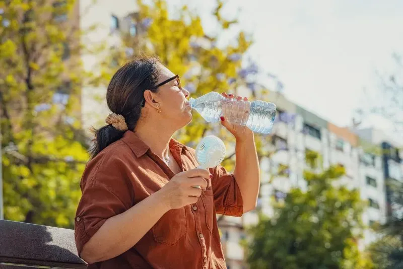 امرأة تشرب الماء في يوم مشمس، تحمل مروحة يدوية، وتظهر أشجار خضراء ومباني في الخلفية، مما يعكس أهمية الترطيب في الطقس الحار.