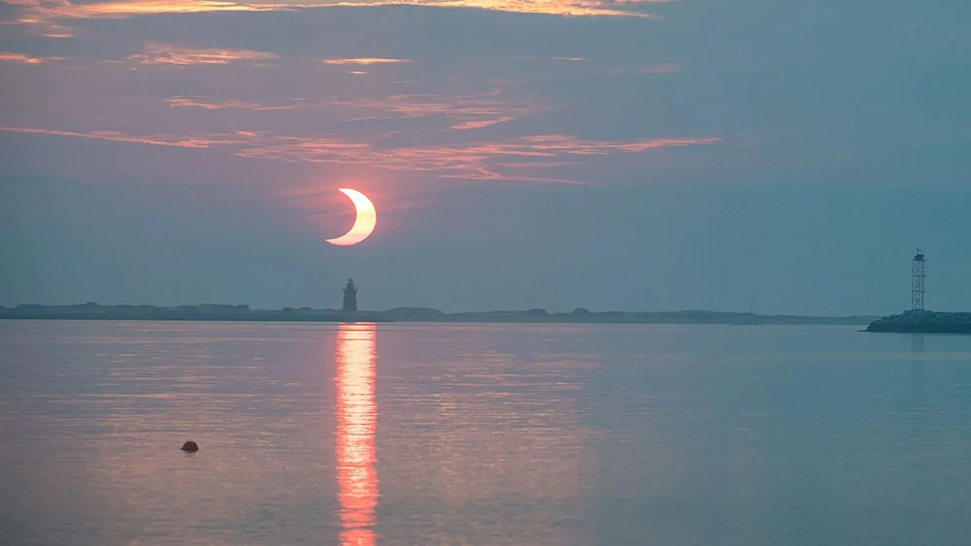 كسوف جزئي للشمس يظهر فيه القمر يحجب جزءًا من الشمس، مع انعكاس الضوء على سطح الماء ومنارة في الخلفية.