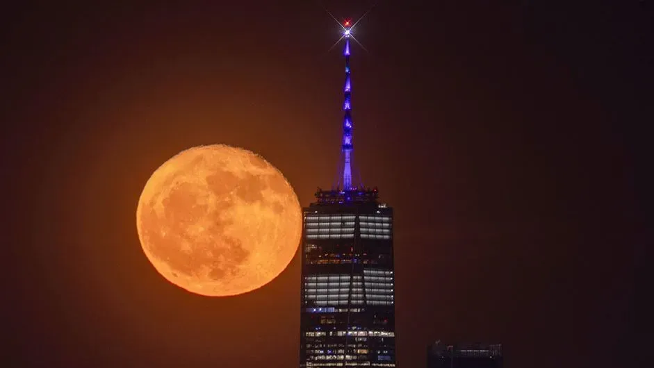 قمر مايو المكتمل يظهر بلون برتقالي ساطع خلف برج في مدينة نيويورك، مع أضواء زرقاء تزين قمة البرج في السماء المظلمة.