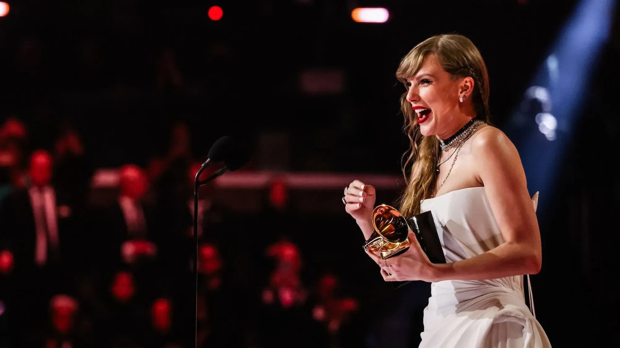 تظهر الصورة فنانة مشهورة تحتفل بفوزها بجائزة جرامي، مرتدية فستاناً أنيقاً وتبتسم بسعادة، مع جمهور خلفها.