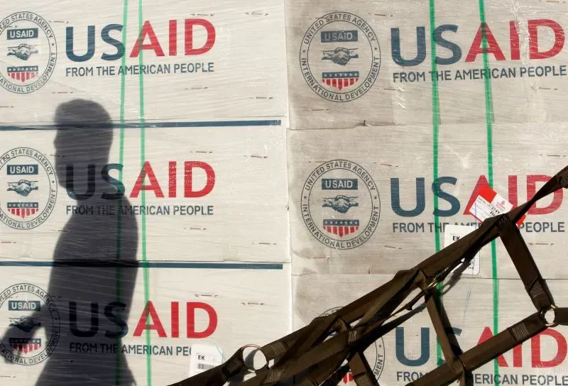 صورة تظهر شخصًا يسير بجانب صناديق تحمل شعار الوكالة الأمريكية للتنمية الدولية (USAID)، مما يعكس تأثير الإدارة الجديدة على القوى العاملة الفيدرالية.