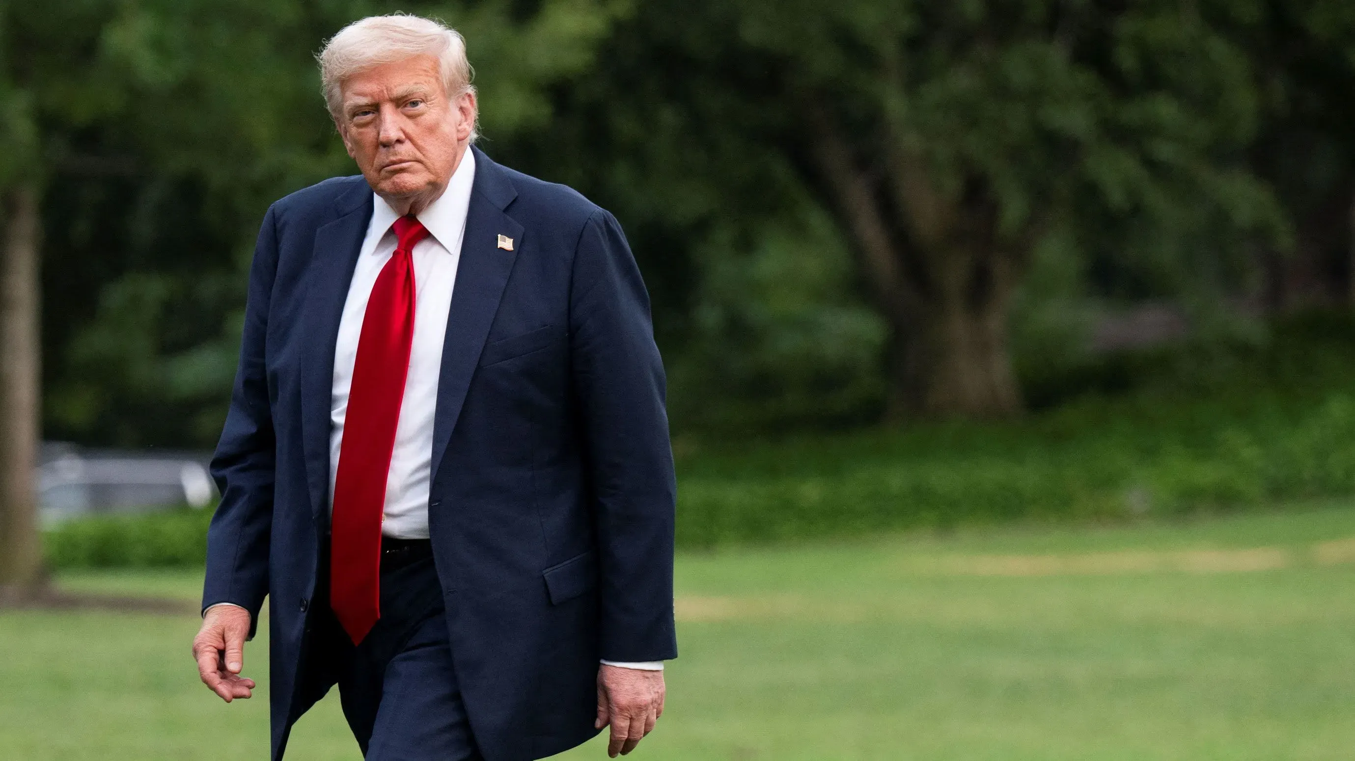 ترامب يسير في حديقة مع تعبير جاد، مرتديًا بدلة سوداء وقميصًا أبيض، ويعكس تأثير سياسته التجارية على الأسواق.
