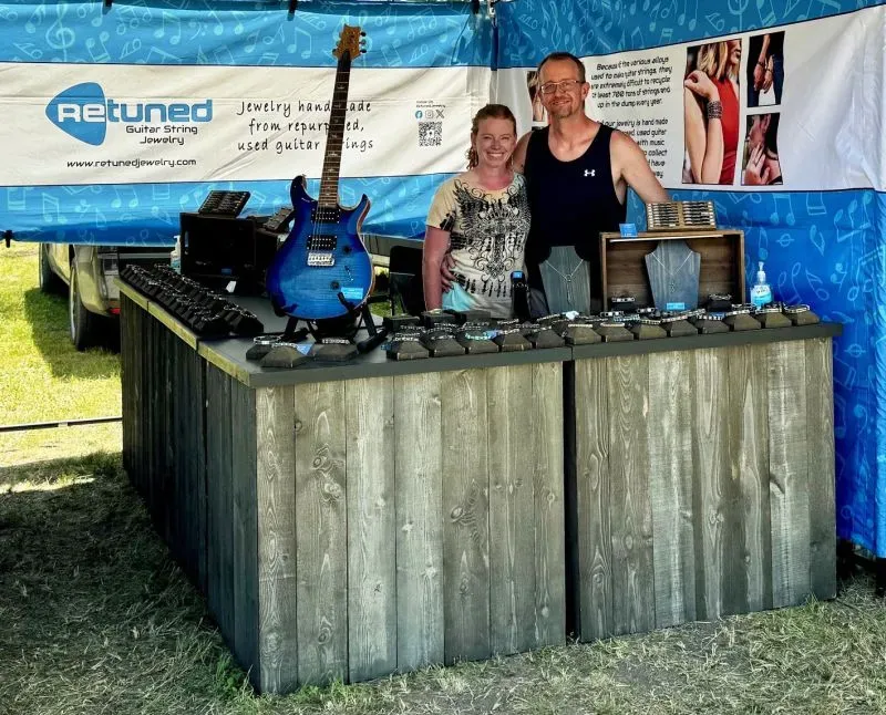 زوجان يقفان أمام طاولة عرض مجوهرات مصنوعة من أوتار الغيتار المعاد تدويرها، مع لافتة لمشروعهما "ريتونيد للمجوهرات".