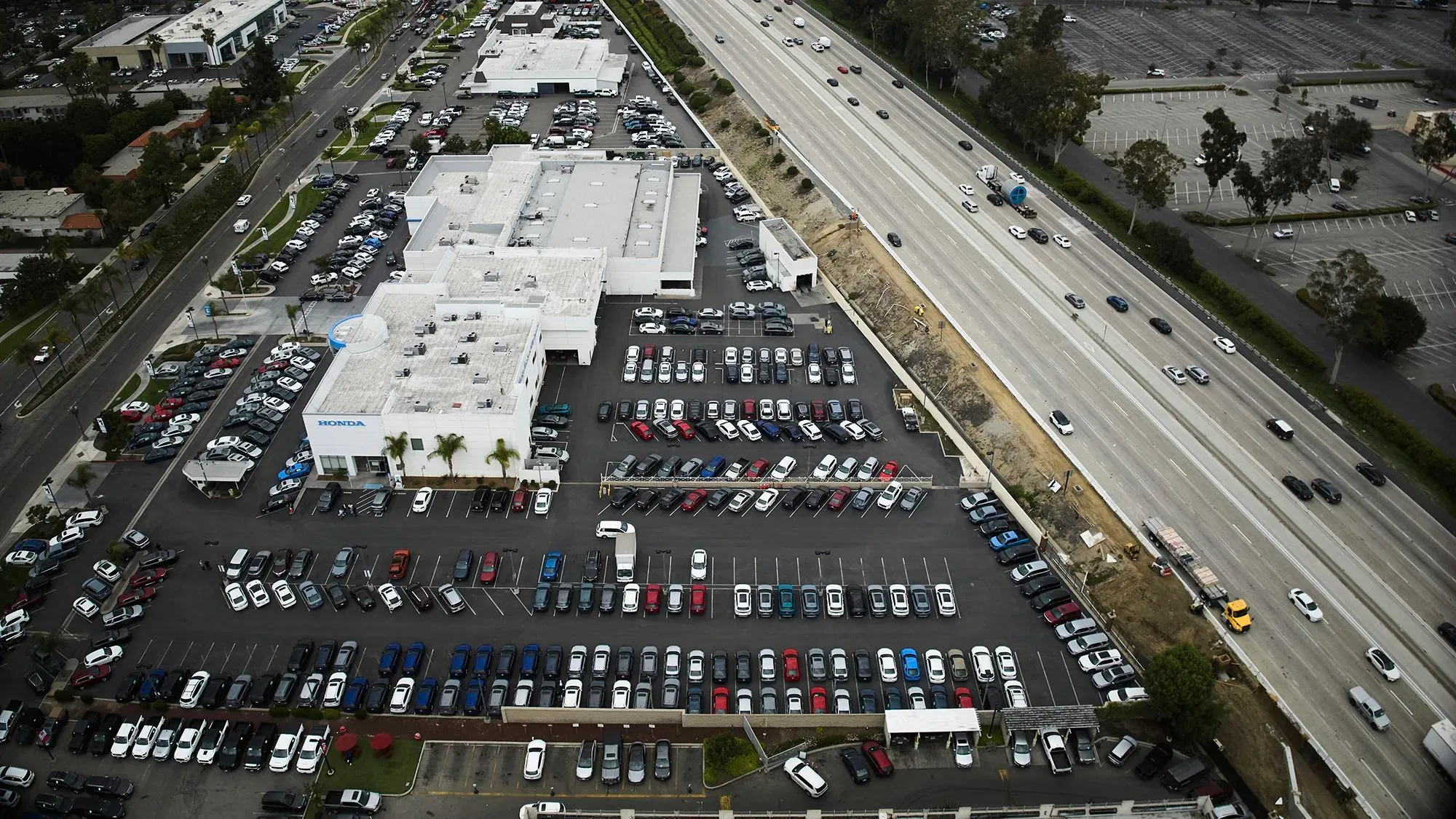 موقف سيارات كبير يضم مجموعة متنوعة من السيارات، مع وجود طريق سريع بجانبه، يعكس تأثير التعريفات الجمركية على أسعار السيارات.