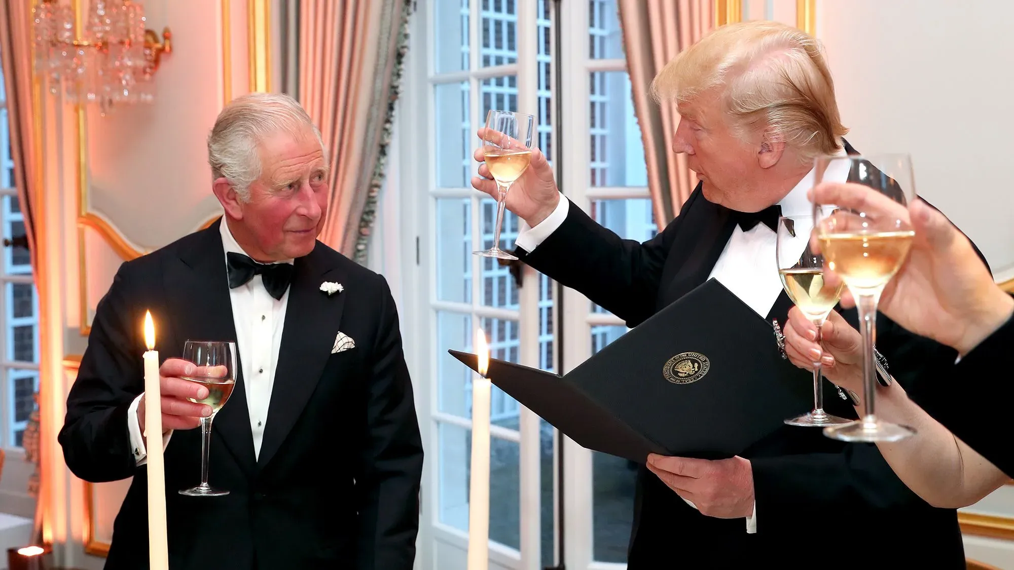 ترامب وشارلز الثالث يتبادلان التحية خلال حفل رسمي، مع وجود شمعة مضاءة وكؤوس مشروبات في الأيدي، مما يعكس العلاقات الدبلوماسية بين الولايات المتحدة والمملكة المتحدة.