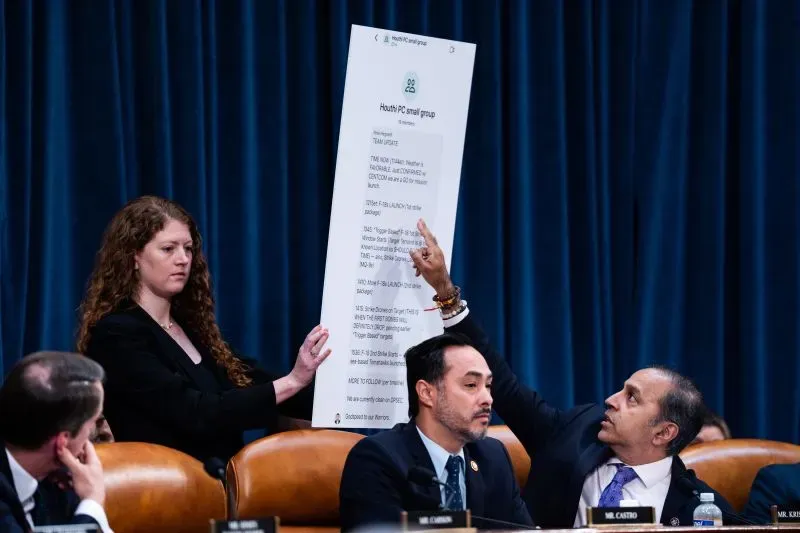 امرأة تحمل لوحة كبيرة تحتوي على معلومات بريد إلكتروني خلال جلسة استماع، بينما يجلس ثلاثة رجال في الخلف. تعكس الصورة الفوضى التنظيمية في الإدارة.