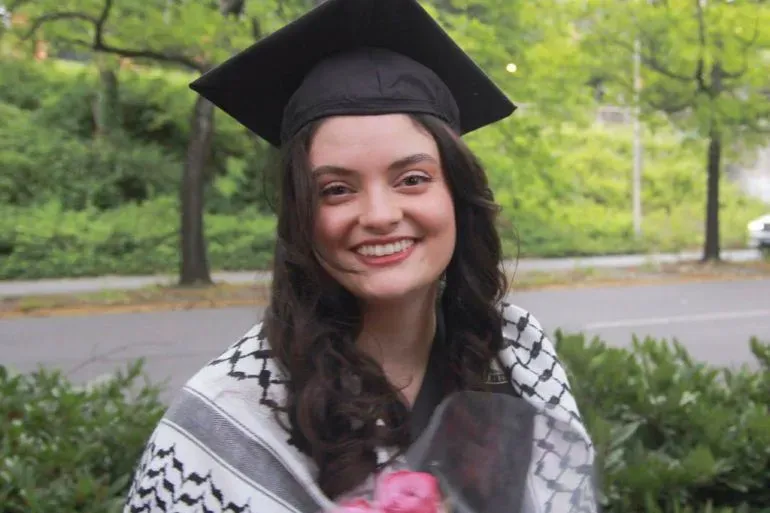 فتاة ترتدي قبعة تخرج وتبتسم، تحمل باقة من الزهور، وتظهر خلفها أشجار خضراء، تعبيرًا عن الفرح والنجاح.