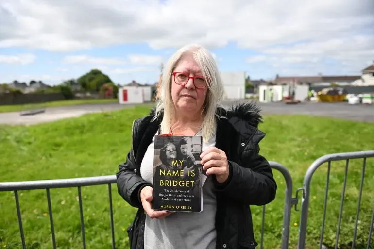 امرأة تحمل كتاباً بعنوان "اسمي بريدجيت" في موقع التنقيب عن مقابر جماعية لأطفال في توام، أيرلندا، حيث تم اكتشاف رفات حوالي 800 رضيع.