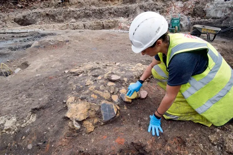 عالمة آثار ترتدي خوذة وقميص أمان، تعمل على تنظيف قطع من الجص في موقع تنقيب في ساوثوارك، حيث تم اكتشاف لوحات جدارية رومانية.