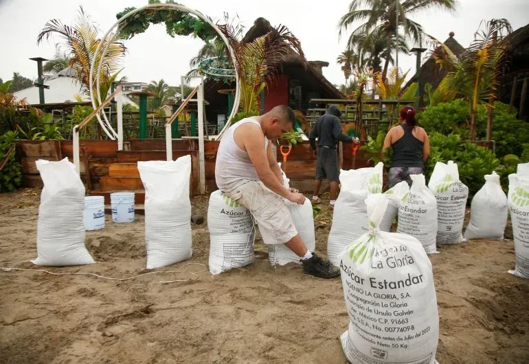 عمال يقومون بتعبئة أكياس الرمل في منتجع شاطئي استعدادًا للإعصار إيريك، مع خلفية من الأشجار والمرافق المتضررة.