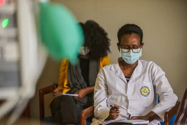 طبيبة إثيوبية ترتدي سترة طبية وقناع وجه، تركز على ملاحظاتها في غرفة انتظار بمستشفى، تعكس تحديات العاملين في القطاع الصحي.