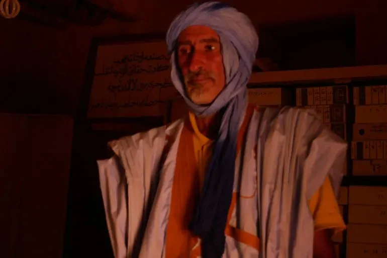 محمد غولام الحبوط في مكتبة تقليدية بشنقيط، يرتدي قفازات بيضاء ويستعرض مخطوطات قديمة، معبراً عن أهمية الحفاظ على التراث الثقافي.