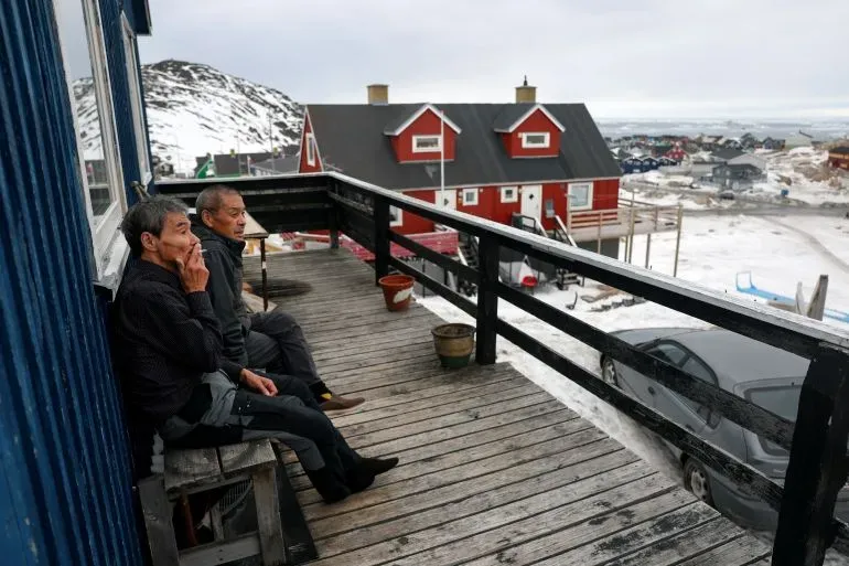 رجلان يجلسان على شرفة خشبية في إيلوليسات، غرينلاند، مع منازل حمراء في الخلفية، يعكسان القلق بشأن تأثيرات الاستحواذ الأمريكي.