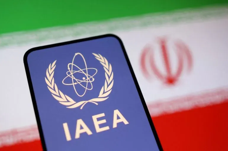 شعار الوكالة الدولية للطاقة الذرية (IAEA) أمام علم إيران، يرمز إلى التوترات حول البرنامج النووي الإيراني وعمليات التفتيش المطلوبة.
