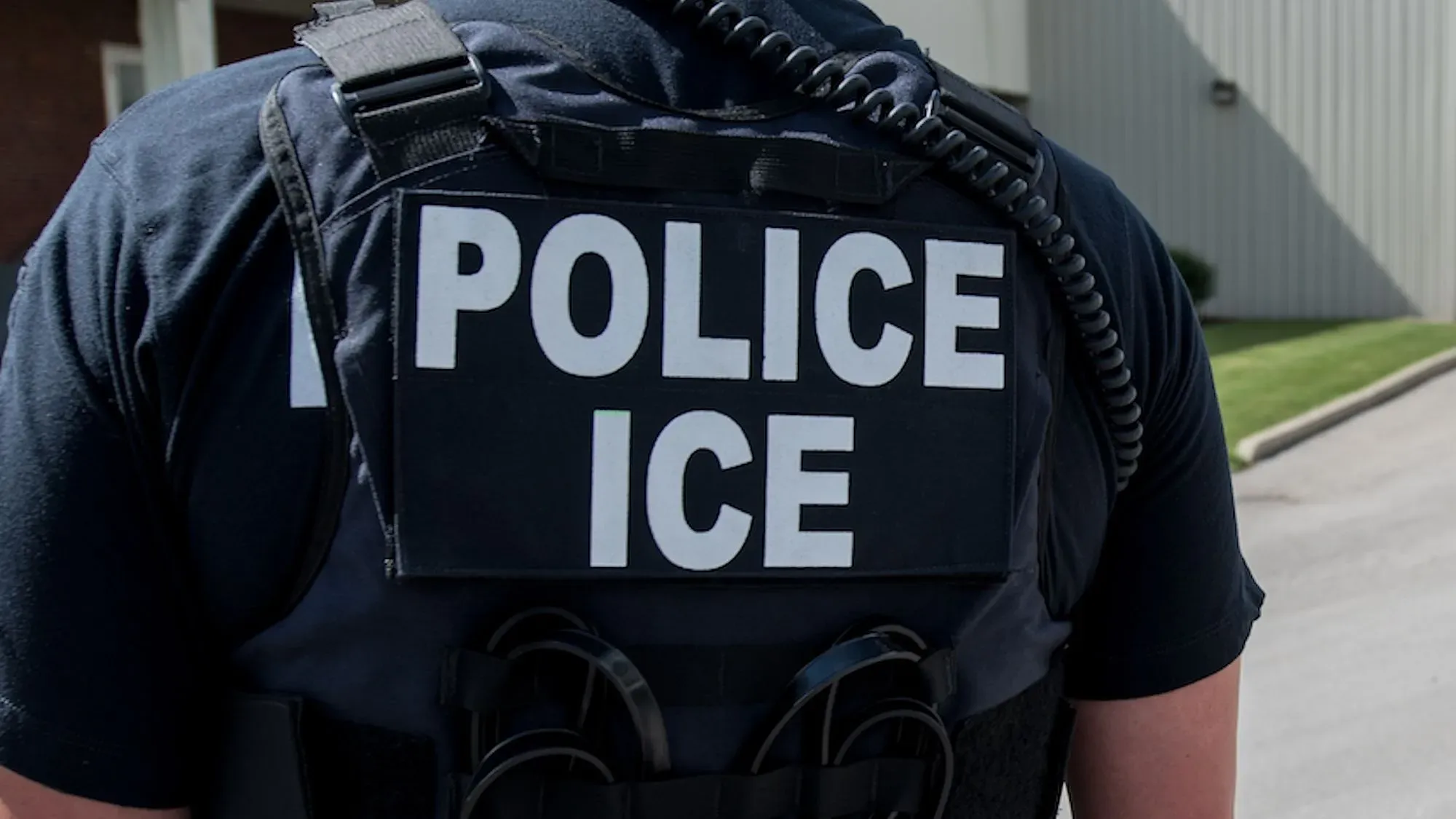 شخص يرتدي سترة سوداء تحمل عبارة "شرطية ICE" مع سماعة أذن، في منطقة بالقرب من مدرسة ابتدائية في شيكاغو.