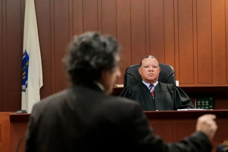 مشهد من قاعة المحكمة في بوسطن، حيث يتحدث محامي أمام القاضي بينما يظهر علم ماساتشوستس خلفهم، في سياق قضية اعتقال مهاجر غير موثق.