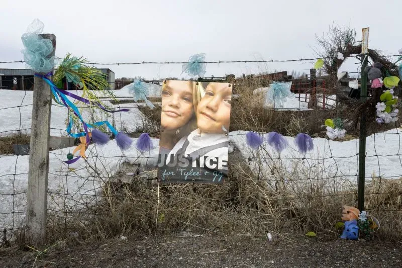 لافتة تحمل صورة لطفلين مع عبارة "العدالة لتايل وتي جي"، محاطة بزينة ملونة على سياج في منطقة ريفية.