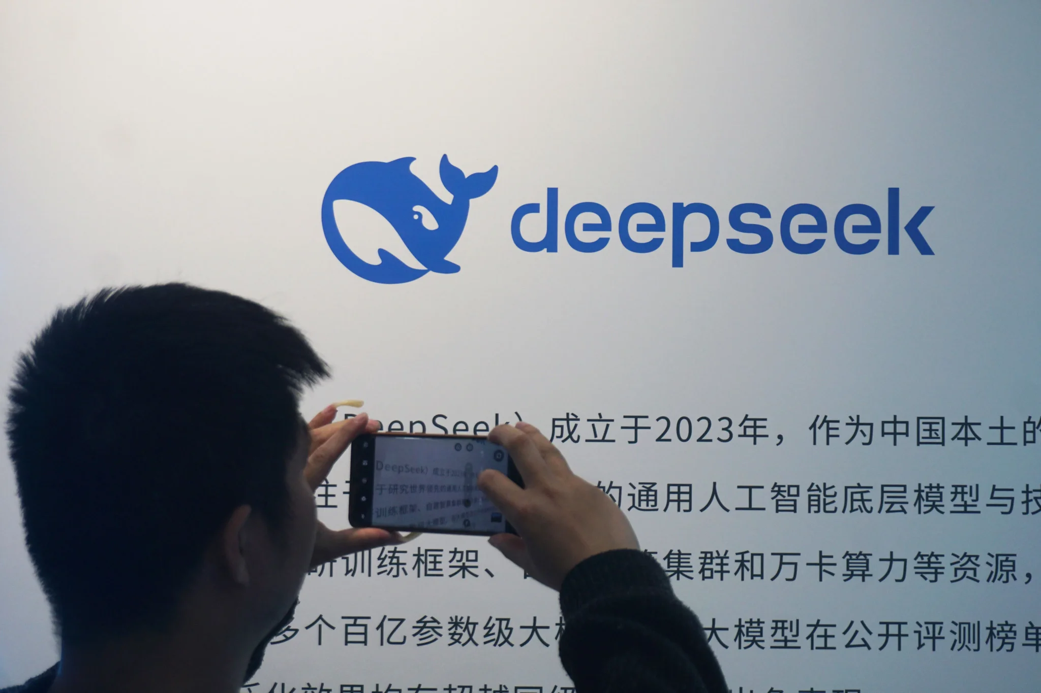 شخص يلتقط صورة بشاشة تعرض شعار DeepSeek ونص صيني، مع التركيز على تطور الذكاء الاصطناعي الصيني وتنافسه مع النماذج الأمريكية.