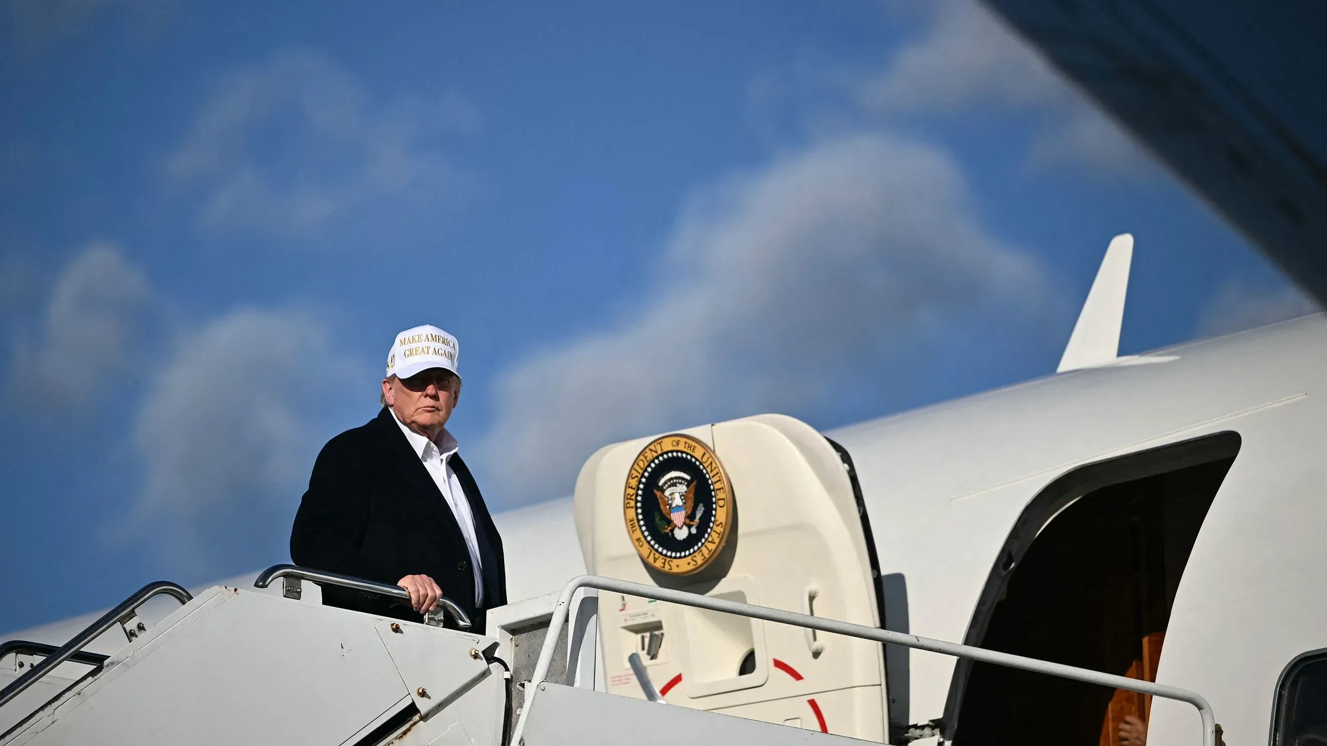 ترامب يرتدي قبعة "اجعل أمريكا عظيمة مرة أخرى" في مطار، بينما يستعد للصعود إلى الطائرة الرئاسية، مع وجود شعار الرئاسة خلفه.