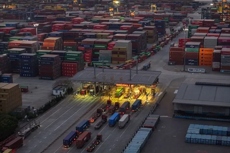 مستودع حاويات كبير يضم شاحنات متوقفة، مع تكدس الحاويات الملونة، يعكس تأثير التجارة العالمية على الاقتصاد.