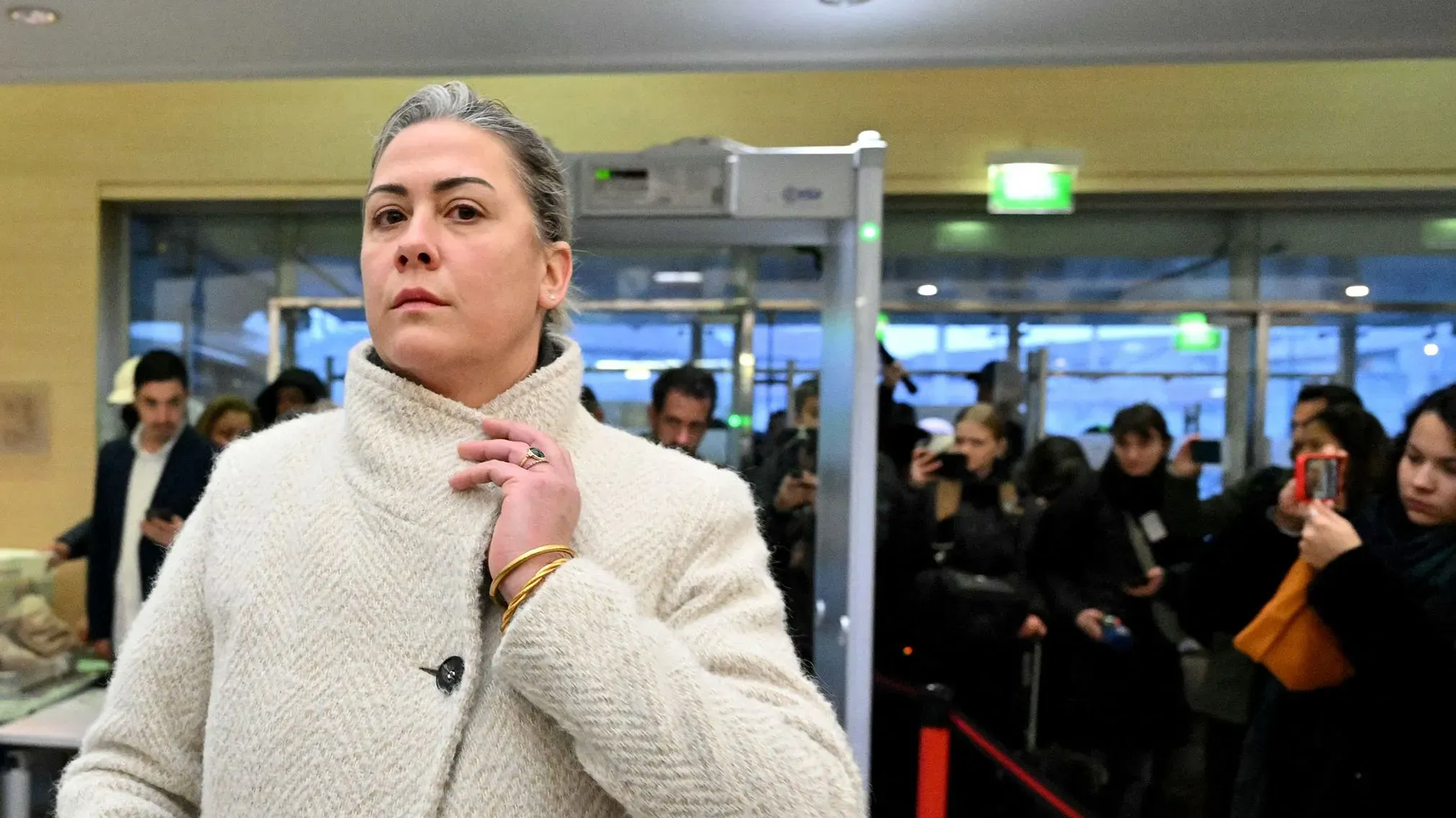 امرأة ترتدي معطفًا أبيض، تدخل مكانًا مزدحمًا، بينما يلتقط الحضور صورًا لها. تعكس تعابير وجهها القوة والإصرار.