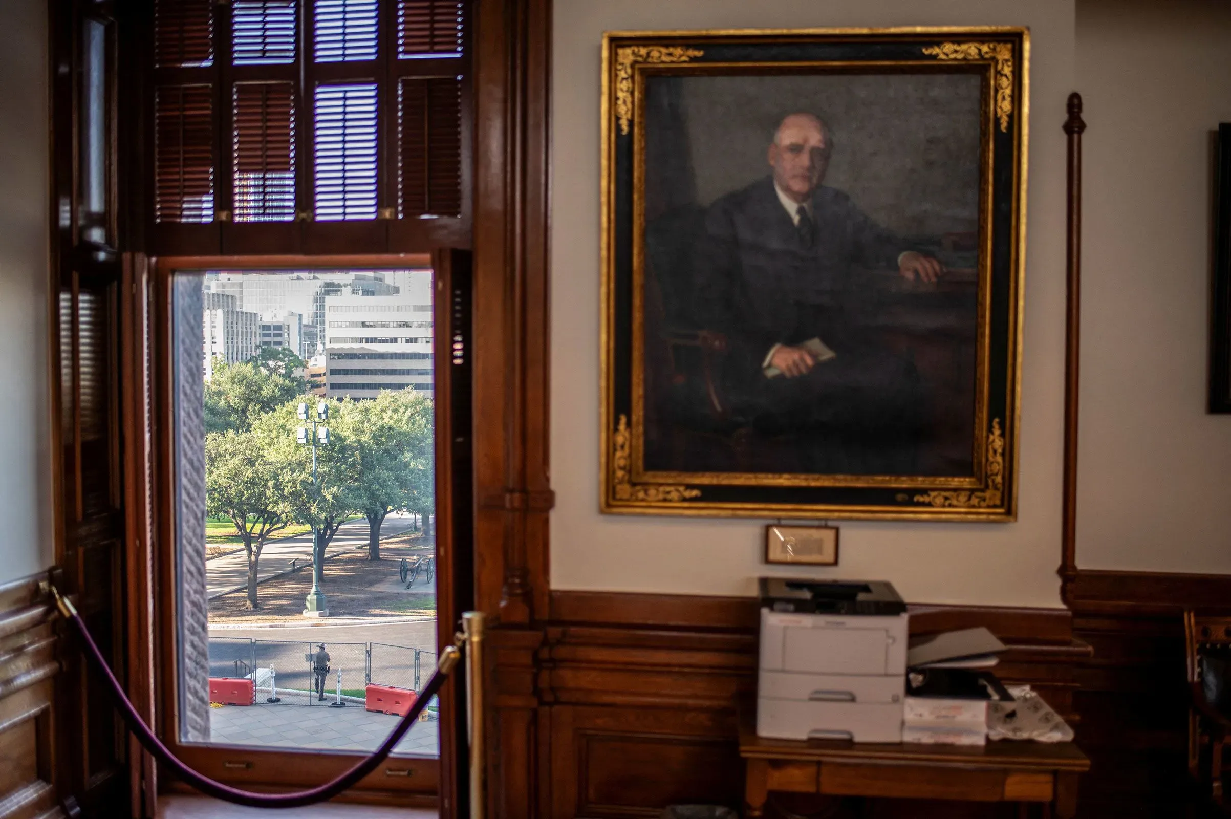 منظر من نافذة مكتب يظهر حديقة خارجية مع أشجار، بالإضافة إلى لوحة فنية لرجل يرتدي بدلة، تعكس أجواء السياسة في تكساس.