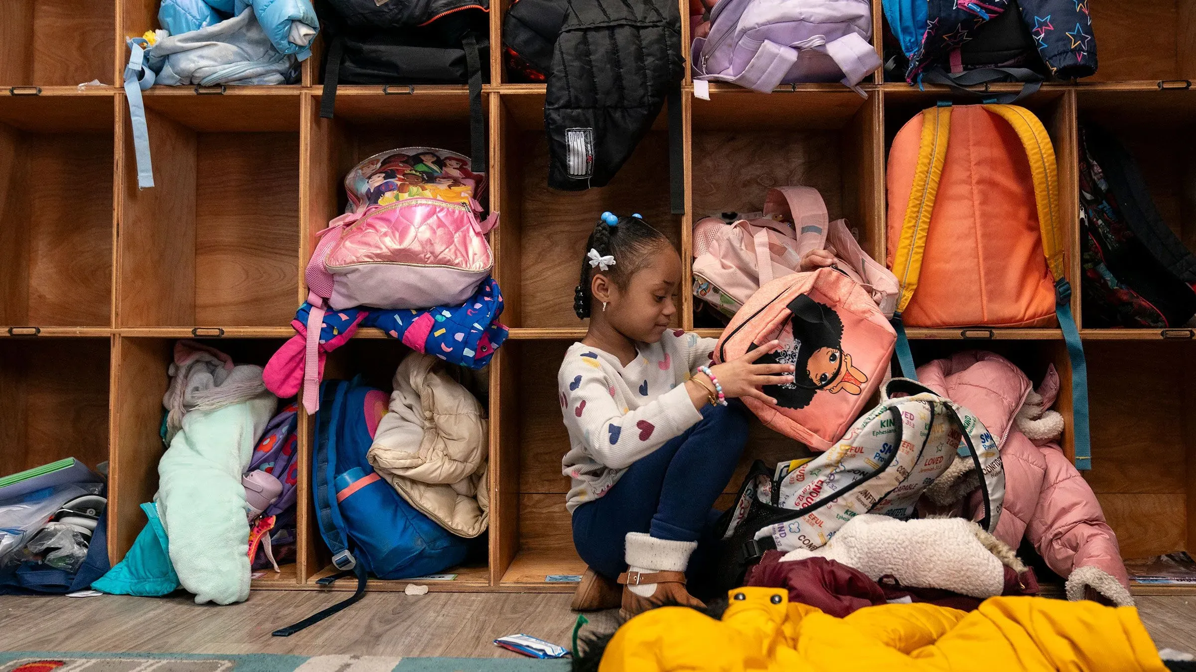 فتاة صغيرة تجلس بين حقائب مدرسية في مركز رعاية نهارية، حيث تعكس الفوضى تحديات رعاية الأطفال في ظل ارتفاع التكاليف.