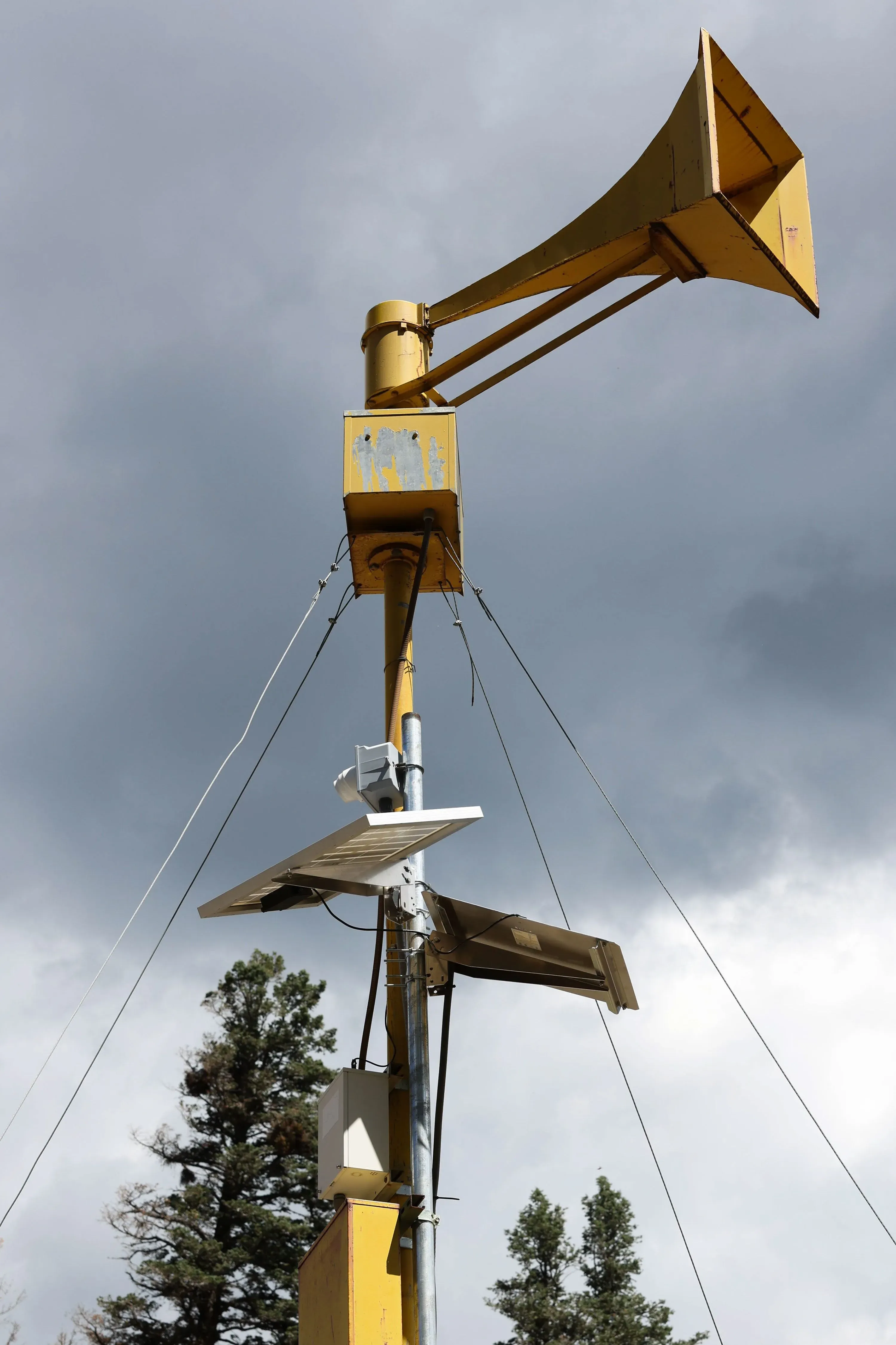 جهاز إنذار أصفر مرتفع يتوسط سماء ملبدة بالغيوم، محاط بأشجار، يُشير إلى التحذيرات من الفيضانات والعواصف في رويدوسو.