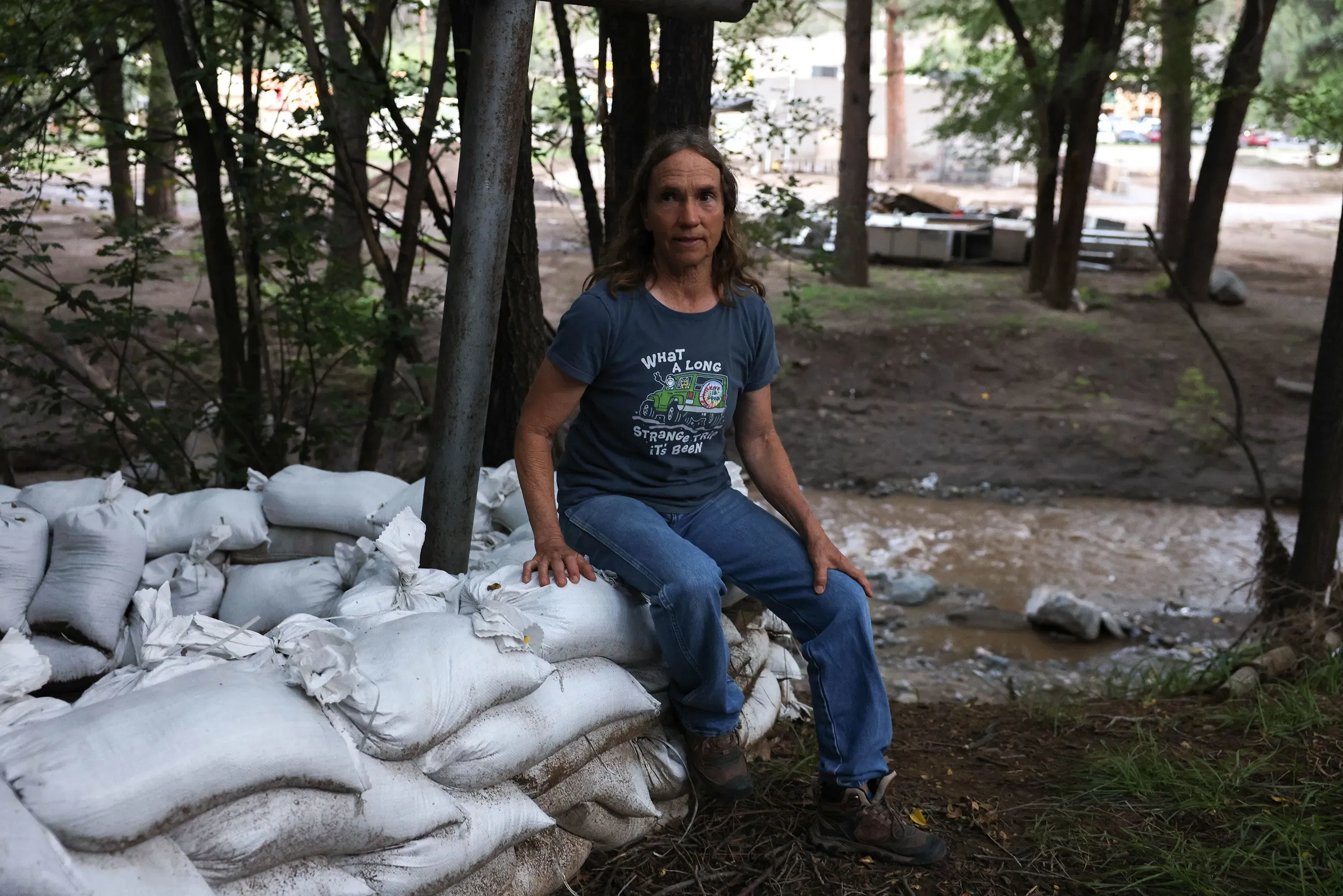 امرأة تجلس على كومة من أكياس الرمل، محاطة بالأشجار، بينما يتدفق الماء من نهر قريب، تعبيرها يوحي بالقلق تجاه الفيضانات المستمرة في رويدوسو.