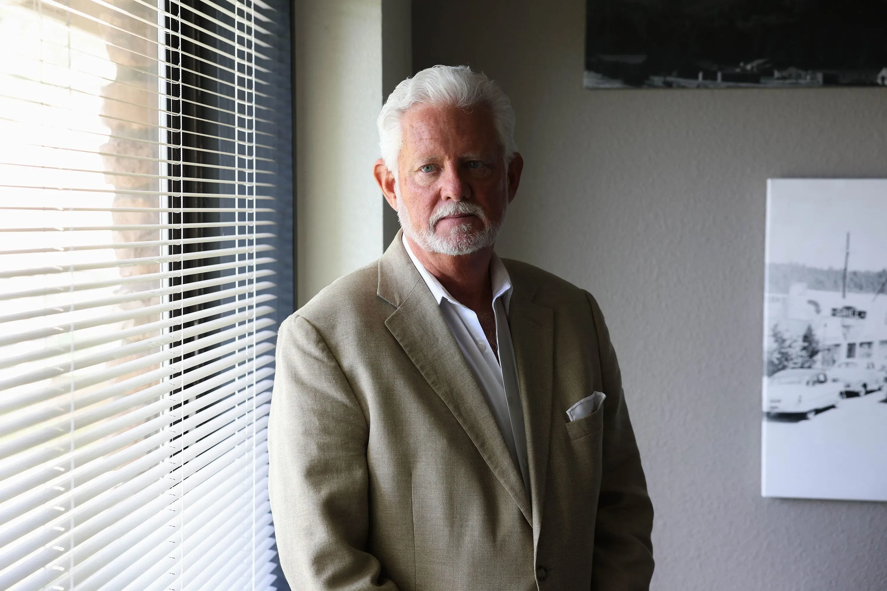 رجل مسن ذو لحية وشعر أبيض يقف بجانب نافذة مع ستائر، يرتدي بدلة رمادية، في خلفية تحتوي على صورة بالأبيض والأسود.