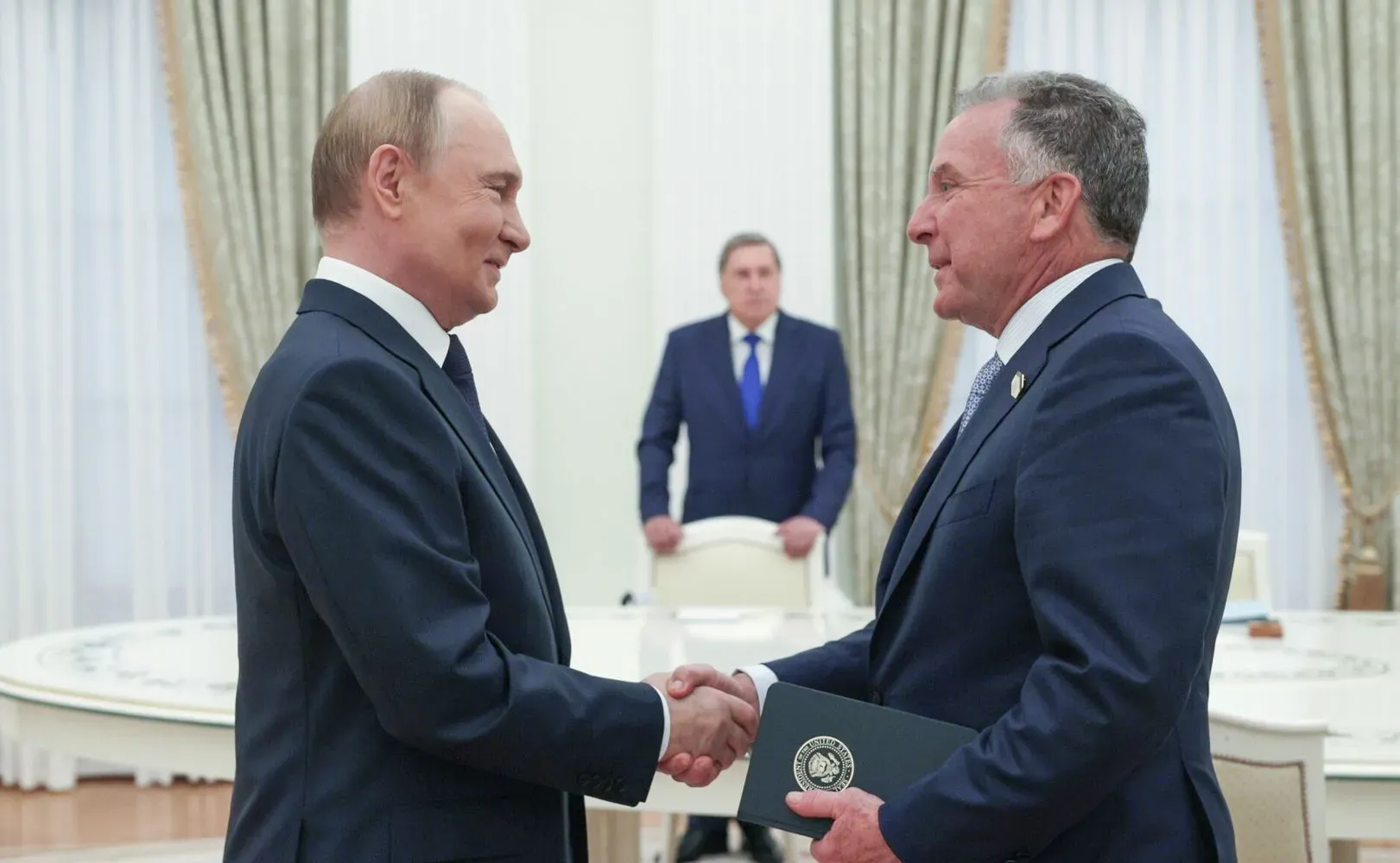 اجتماع بين فلاديمير بوتين ورجل أعمال، مع تعبيرات جدية، في قاعة رسمية. يبرز في الخلفية شخص آخر، مما يضيف سياقًا للحدث.