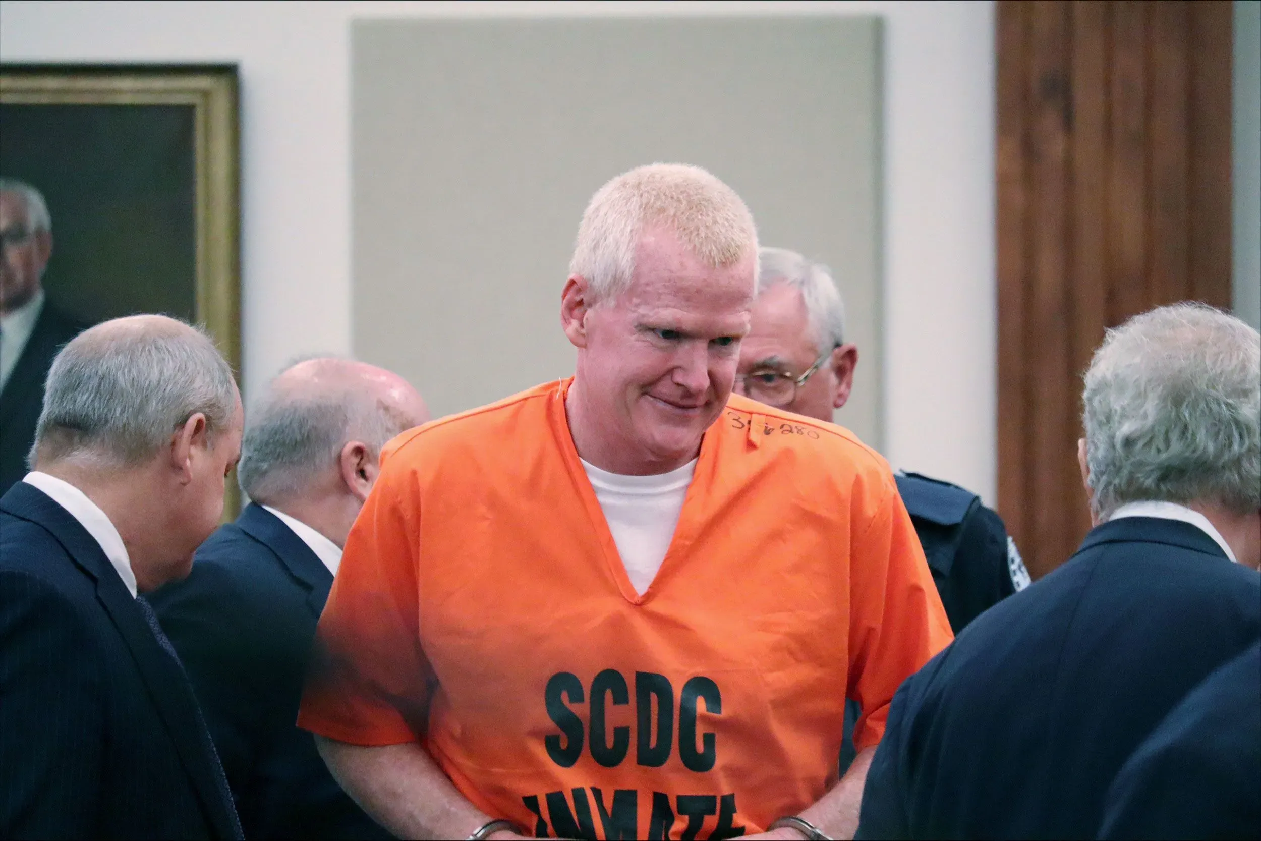 أليكس موردو، المحامي المدان بجريمة القتل، يرتدي زي السجن البرتقالي أثناء جلسة استماع قانونية، محاطًا بمحاميه وأعضاء من المحكمة.