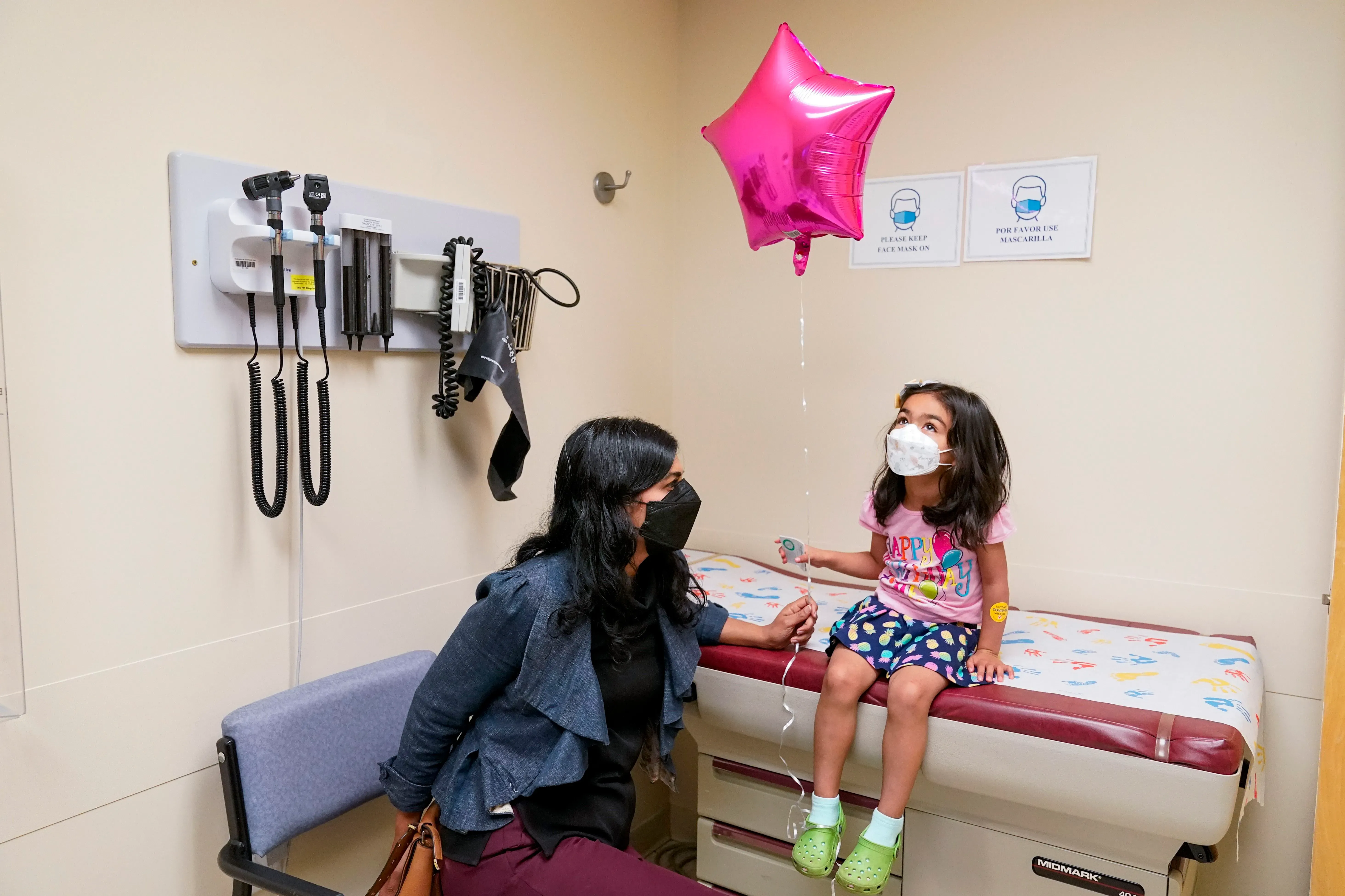 طفلة ترتدي قناع وجه وتجلس على سرير طبي في عيادة، تحمل بالونًا ورديًا، بينما تجلس والدتها بجانبها.
