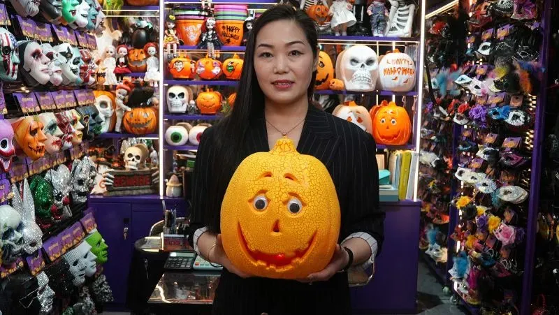 امرأة تحمل قرعًا مبتسمًا في متجر مليء بزينة الهالوين، مع خلفية من الأقنعة والديكورات الملونة.