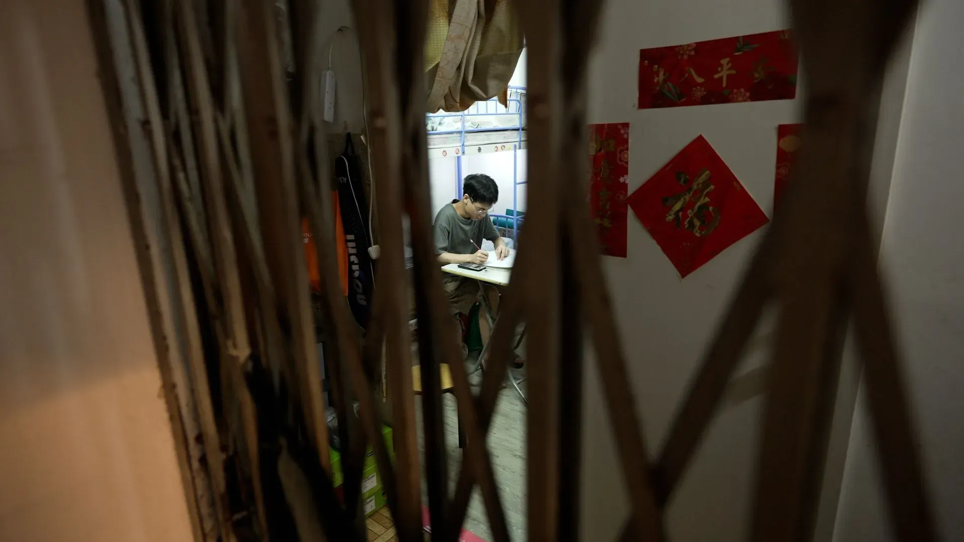 شاب يجلس على طاولة صغيرة في غرفة ضيقة، محاطاً بجدران رقيقة وزينة بسيطة، تعكس ظروف المعيشة الصعبة في هونغ كونغ.