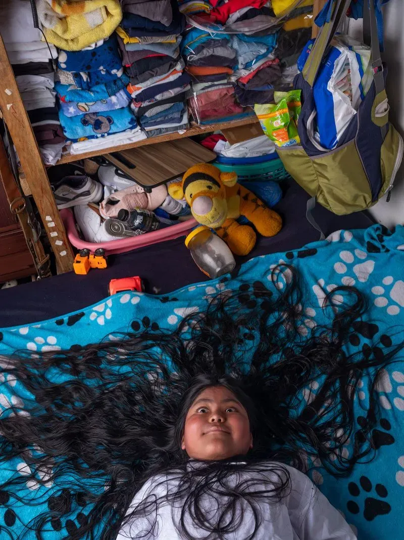 صورة لفتاة صغيرة مستلقية على سرير، شعرها الطويل منتشر حولها. خلفية الغرفة تحتوي على أكوام من الملابس والألعاب، مما يعكس بيئة منزلية.
