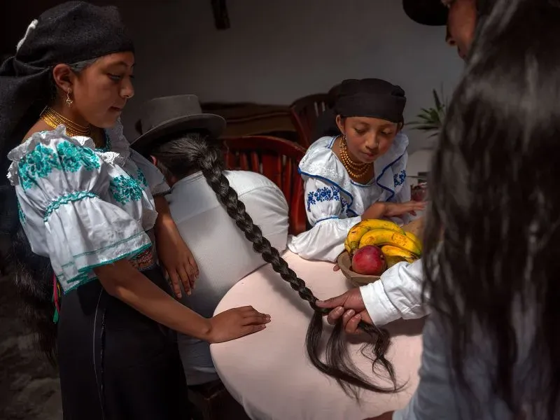 شقيقتان من مجتمع كيشوا ترتديان ملابس تقليدية، بينما يقوم والدهما بتجديل شعر إحداهما، مع وجود فواكه على الطاولة.