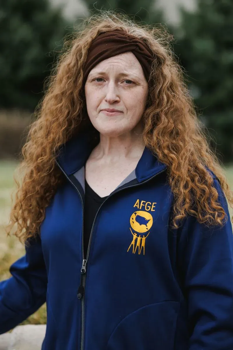 امرأة ذات شعر مجعد طويل ترتدي سترة زرقاء تحمل شعار AFGE، تبدو جادة وتعبر عن القلق بشأن تأثير تسريح العمال الفيدراليين في كانساس سيتي.