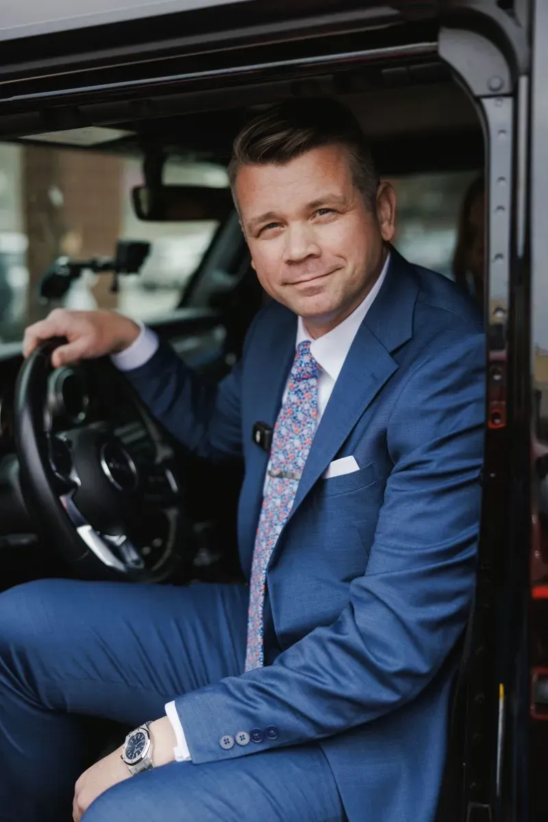 رجل يرتدي بدلة زرقاء يجلس في سيارة، مبتسمًا، مما يعكس حالة القلق في مدينة كانساس سيتي بسبب تسريحات العمال الفيدراليين.