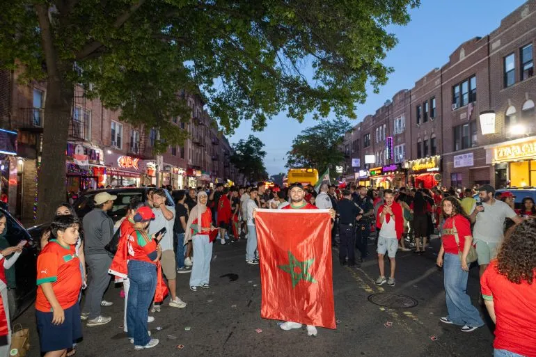 احتفالات حماسية في حي Astoria بنيويورك، حيث يحمل مشجعون مغاربة العلم الوطني وسط أجواء من الفرح بعد تأهل المغرب لكأس العالم.