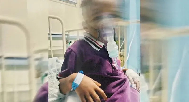 طفل يجلس في مستشفى، يرتدي قميصًا بنفسجيًا، ويستخدم جهاز تنفس. يظهر على يده سوار طبي، مما يشير إلى حالته الصحية.